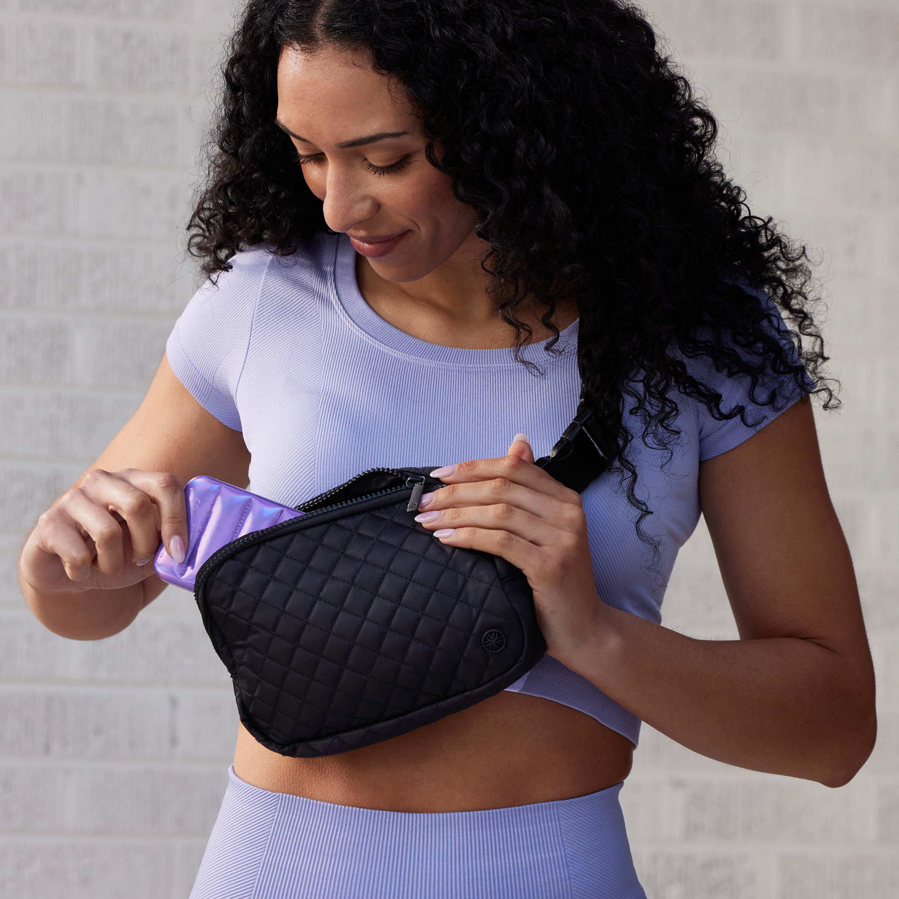 Person standing outside wearing the Sidekick Waist Pack - Quilted XL as a crossbody with the zippered pouch up and pulling out content 
